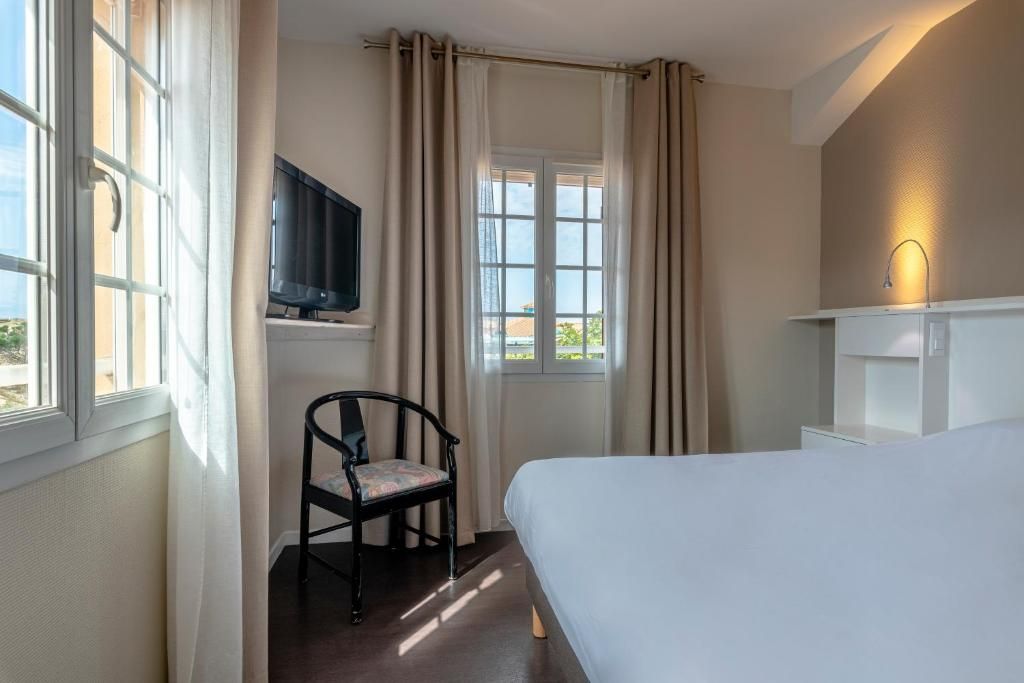Habitación de hotel con cama, una silla, televisor, ventana con cortinas y una lámpara de pared.