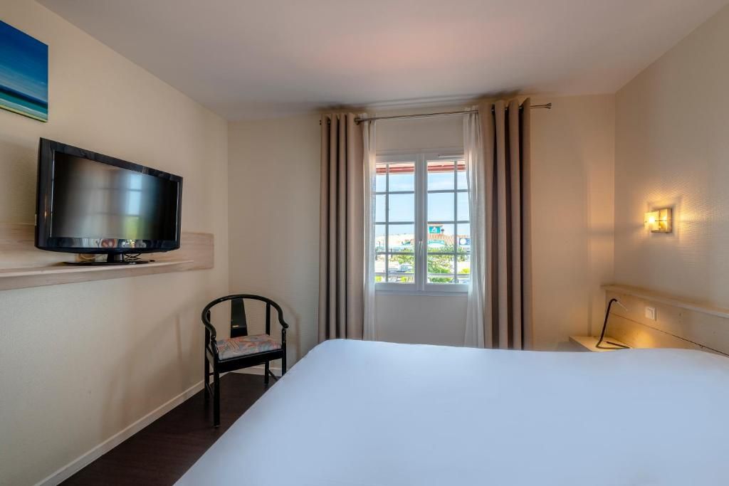 Une chambre d’hôtel avec un lit, un téléviseur posé sur un meuble, une chaise, une fenêtre avec des rideaux et un luminaire fixé au mur.