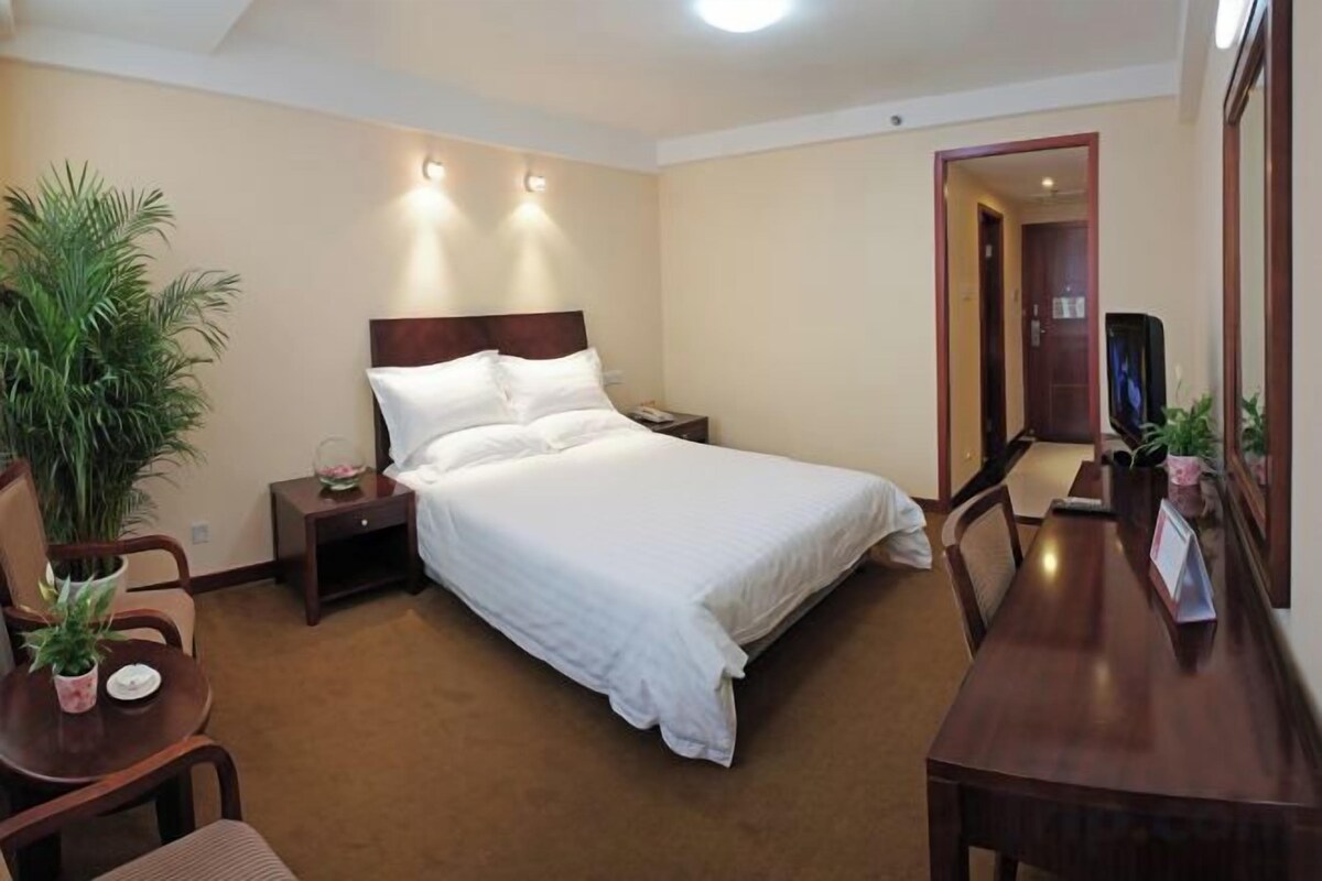 A hotel room with a bed, bedside table, lamp, chair, and a small table with a plant.