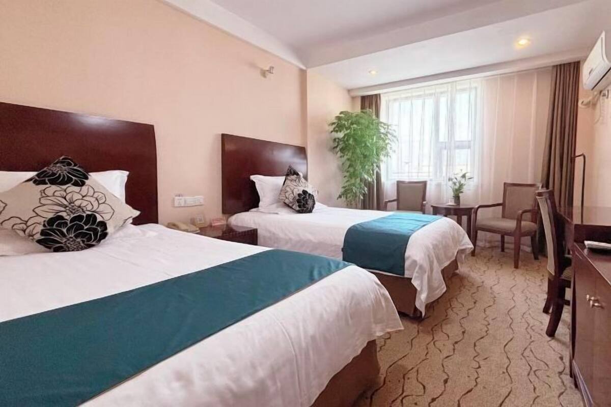 Ein Hotelzimmer mit zwei Betten, einem Schreibtisch, einem Stuhl, einer Zimmerpflanze und einem Fenster mit Vorhängen.