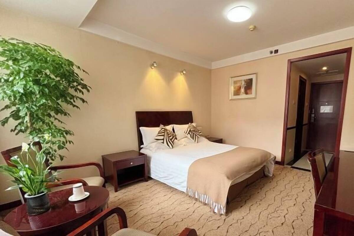 Ein Hotelzimmer mit Bett, Nachttischen, einer Zimmerpflanze, einem Schreibtisch und einem Sessel.