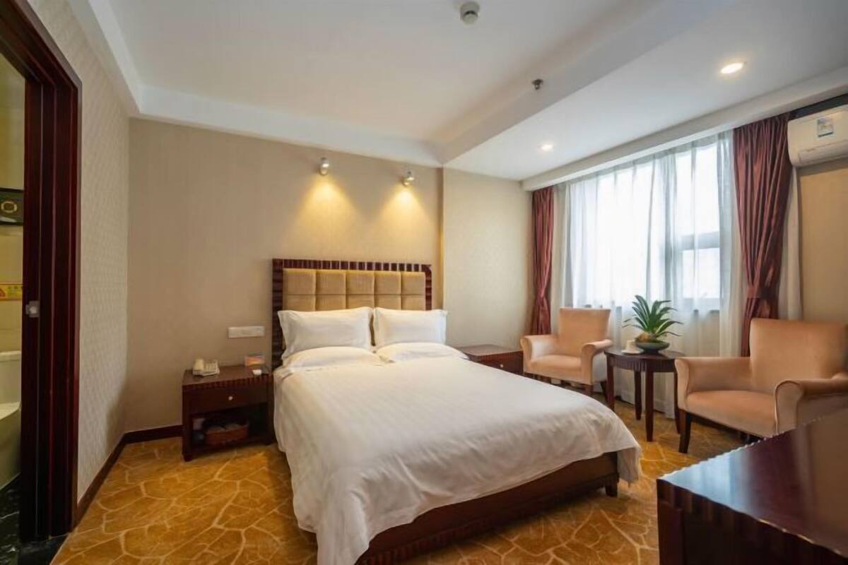 Ein Hotelzimmer mit einem großen Bett, einer Sitzecke mit Sesseln, einem kleinen Tisch mit Pflanze und einem Fenster mit Vorhängen.