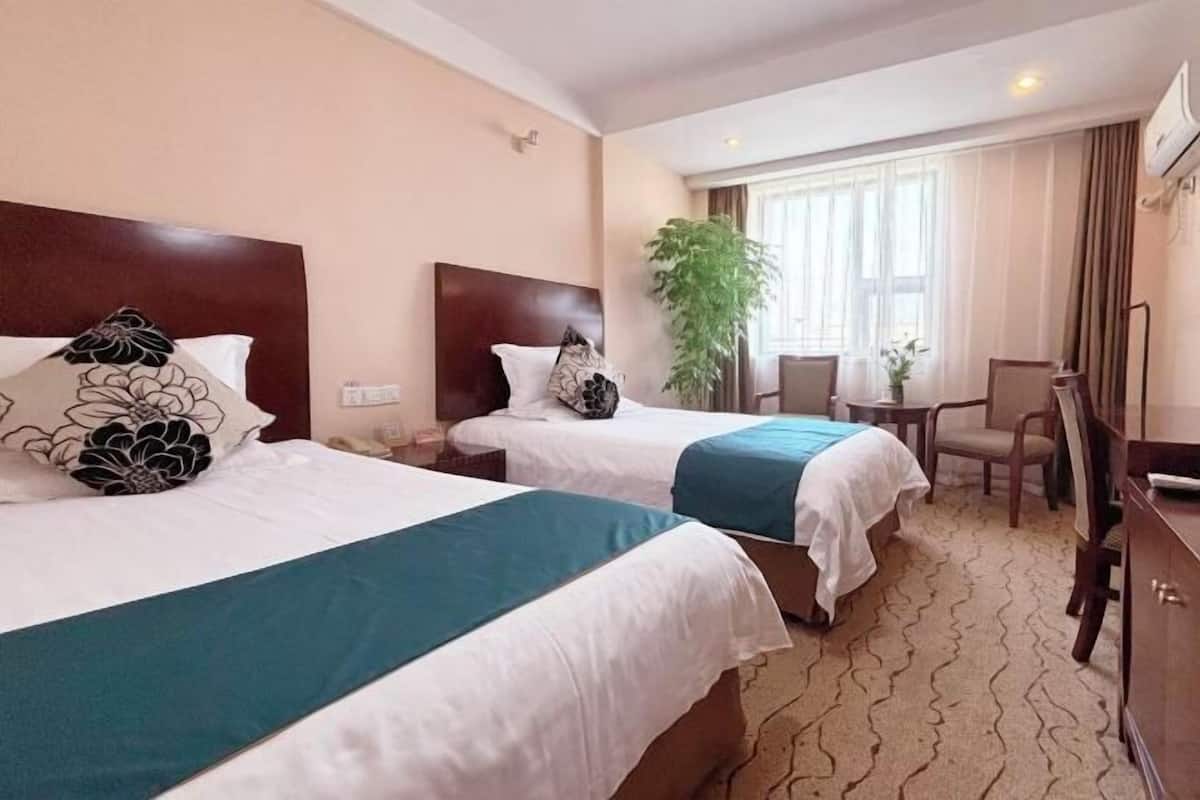 A hotel room with two beds, a desk, a chair, a potted plant, and a window with curtains.