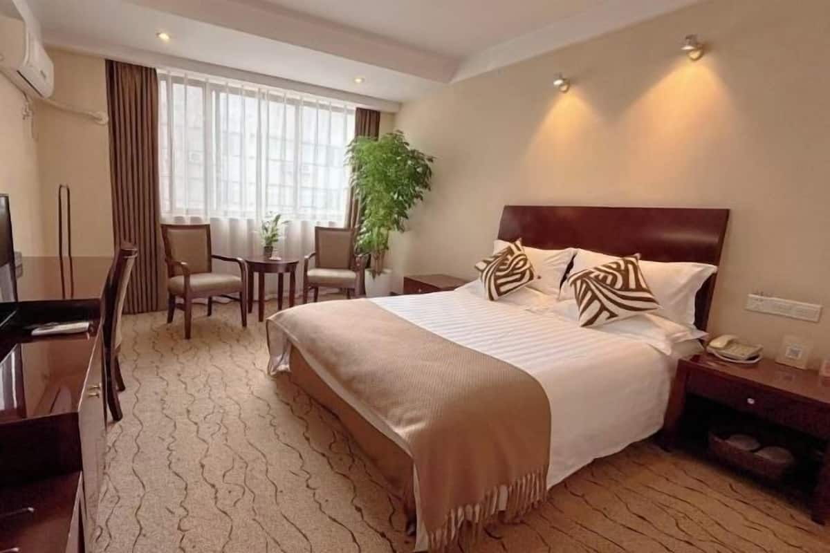 A hotel room with a large bed, a desk, and a chair. There is a plant, a window with curtains, and a wall-mounted phone.