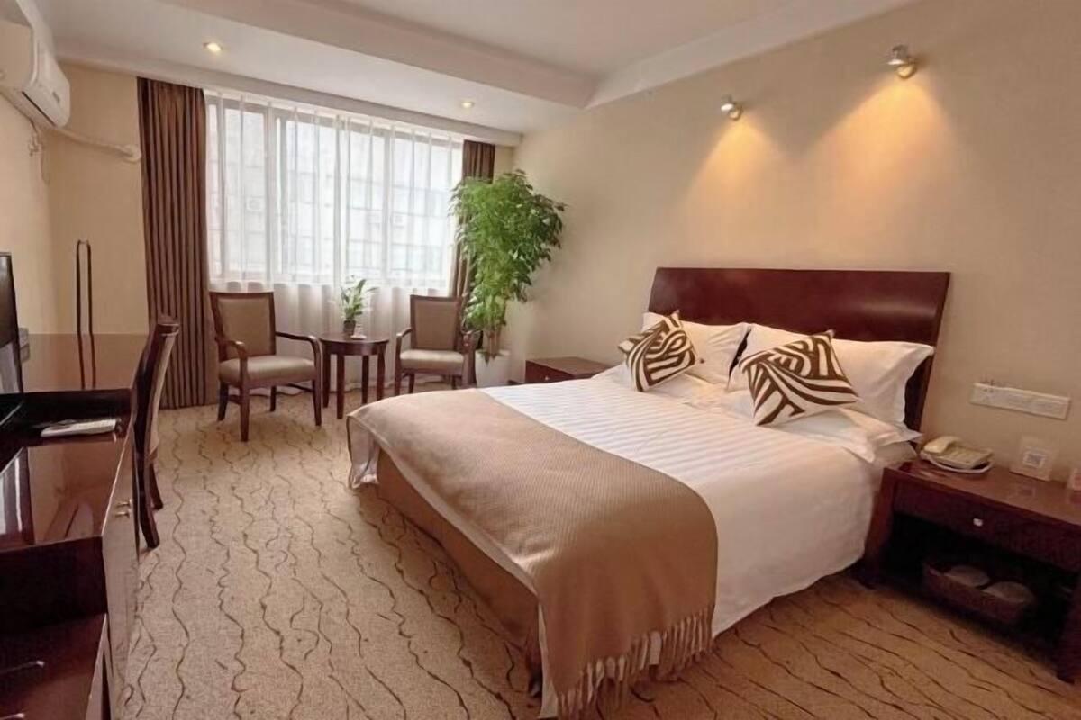 Ein Hotelzimmer mit einem großen Bett, einem Schreibtisch und einem Stuhl. Eine Pflanze, ein Fenster mit Vorhängen und ein an der Wand befestigtes Telefon sind ebenfalls vorhanden.