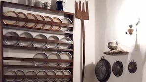 A rustic kitchen with a wooden shelf holding multiple plates, a large fork, and various pots and pans on the wall. - Bendeni Cottage (Chania)