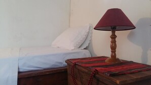 A single bed with a white sheet and a red and black striped blanket, a wooden nightstand with a lamp, and a white wall. - Bendeni Cottage (Chania)
