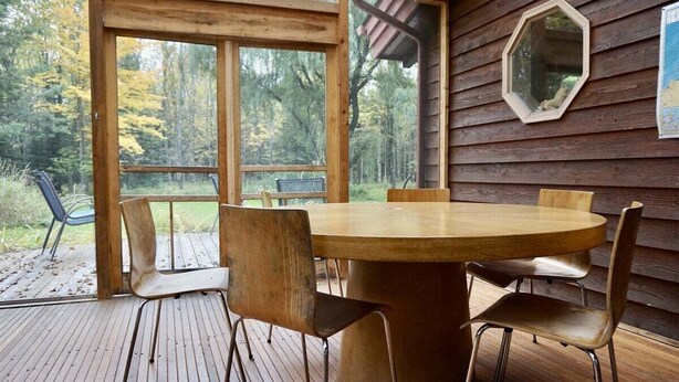 Une terrasse en bois avec une table ronde et des chaises, un miroir hexagonal accroché au mur et une vue sur les arbres à travers de grandes fenêtres.