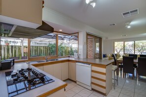 A modern kitchen with a gas stove, dishwasher, and a dining area with a table and chairs. - SHORE THING - FAMILY BEACH RETREAT  (Tootgarook)