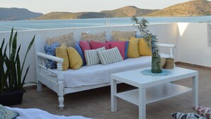 A terrace with a white sofa and table, a potted plant, and a view of the sea and hills. - Akti Posidonos (Agios Nikolaos)