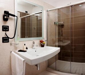 Un baño con un lavamanos blanco, un espejo, un secador de pelo, una ducha con puerta de vidrio y una toalla.