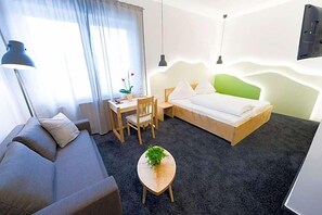 A modern hotel room with a bed, a sofa, a desk, and a small table with a plant.