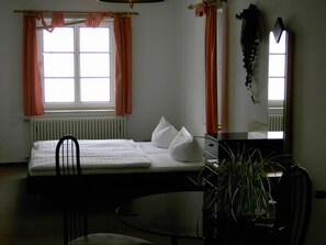 A hotel room with a bed, a desk, a chair, a radiator, and a window with curtains. - Landhotel Am Fuchsbach (Berga/Elster)