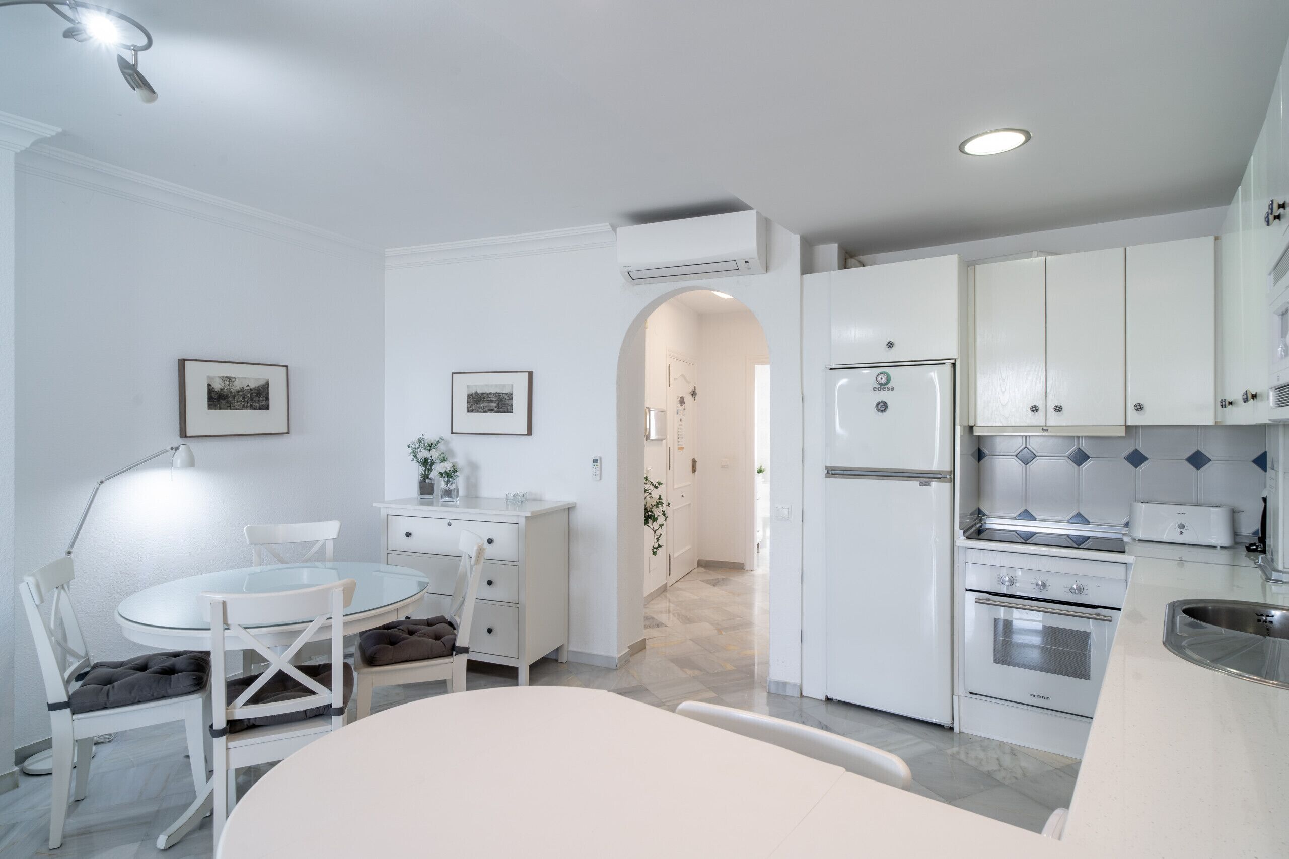 Una cocina moderna con armarios blancos, horno empotrado y un comedor con mesa y sillas de vidrio.