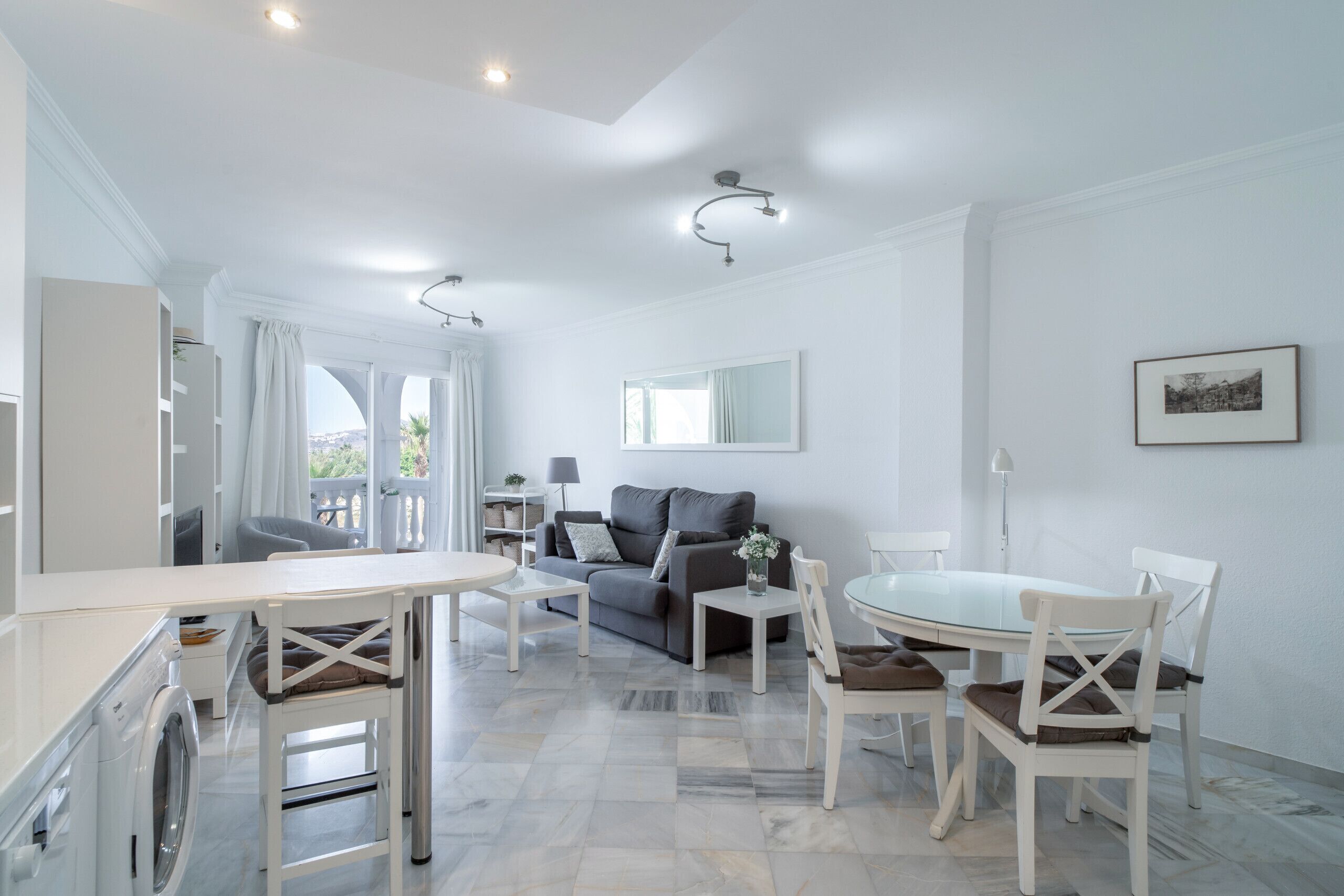 A modern living room with a sofa, coffee table, and dining table. There is a washing machine in the corner.