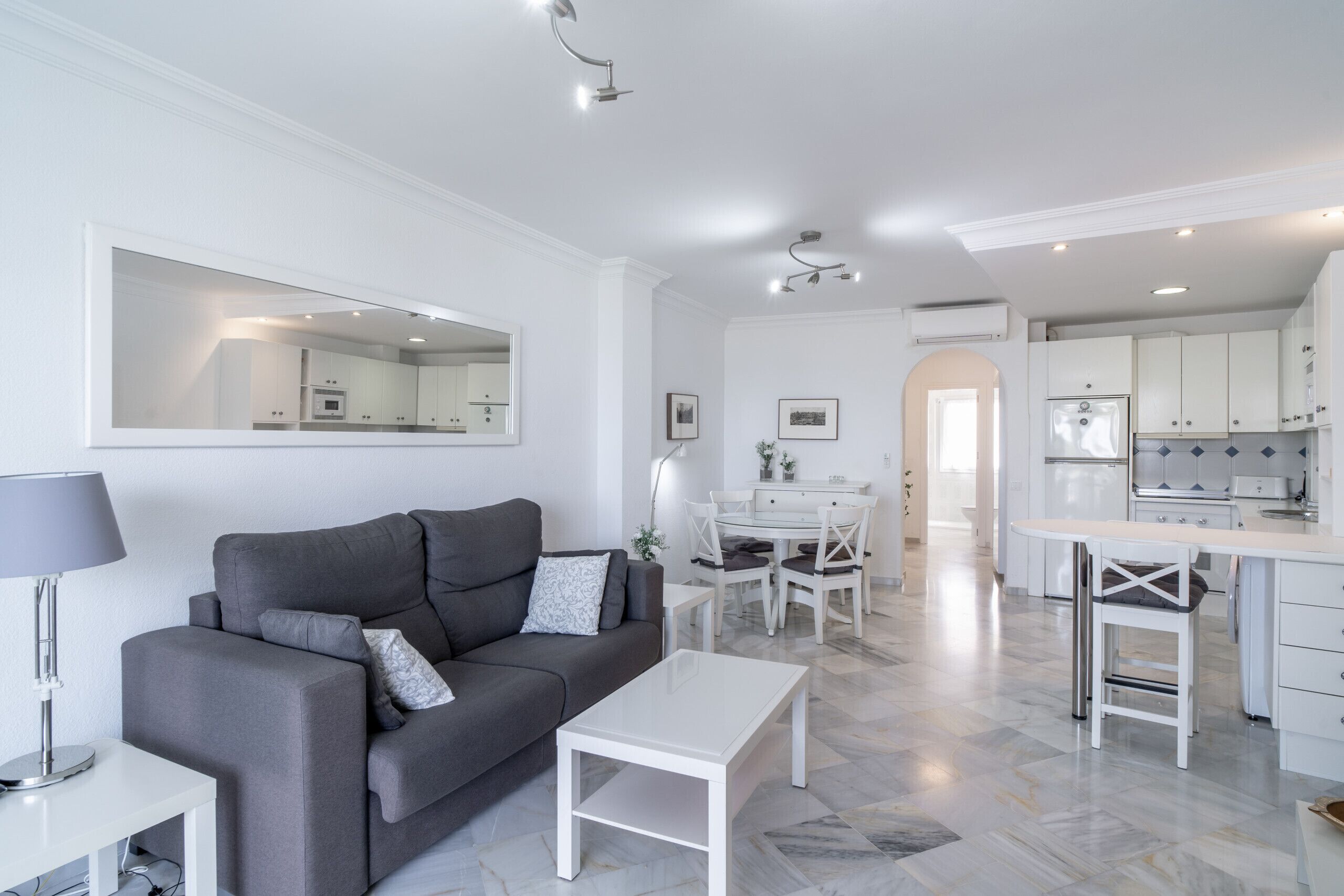 Ein modernes Wohnzimmer mit einem grauen Sofa, einem weißen Couchtisch und einem Essbereich mit weißem Tisch und Stühlen.