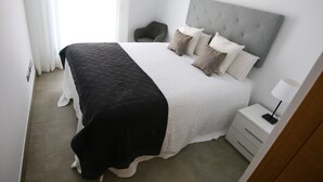 Een netjes opgemaakt bed met een grijze hoofdbord, een nachtkastje met een lamp, en een stoel in een moderne, minimalistische slaapkamer.