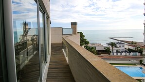 Een balkon met uitzicht op zee, een zwembad en een modern gebouw met een glazen gevel.