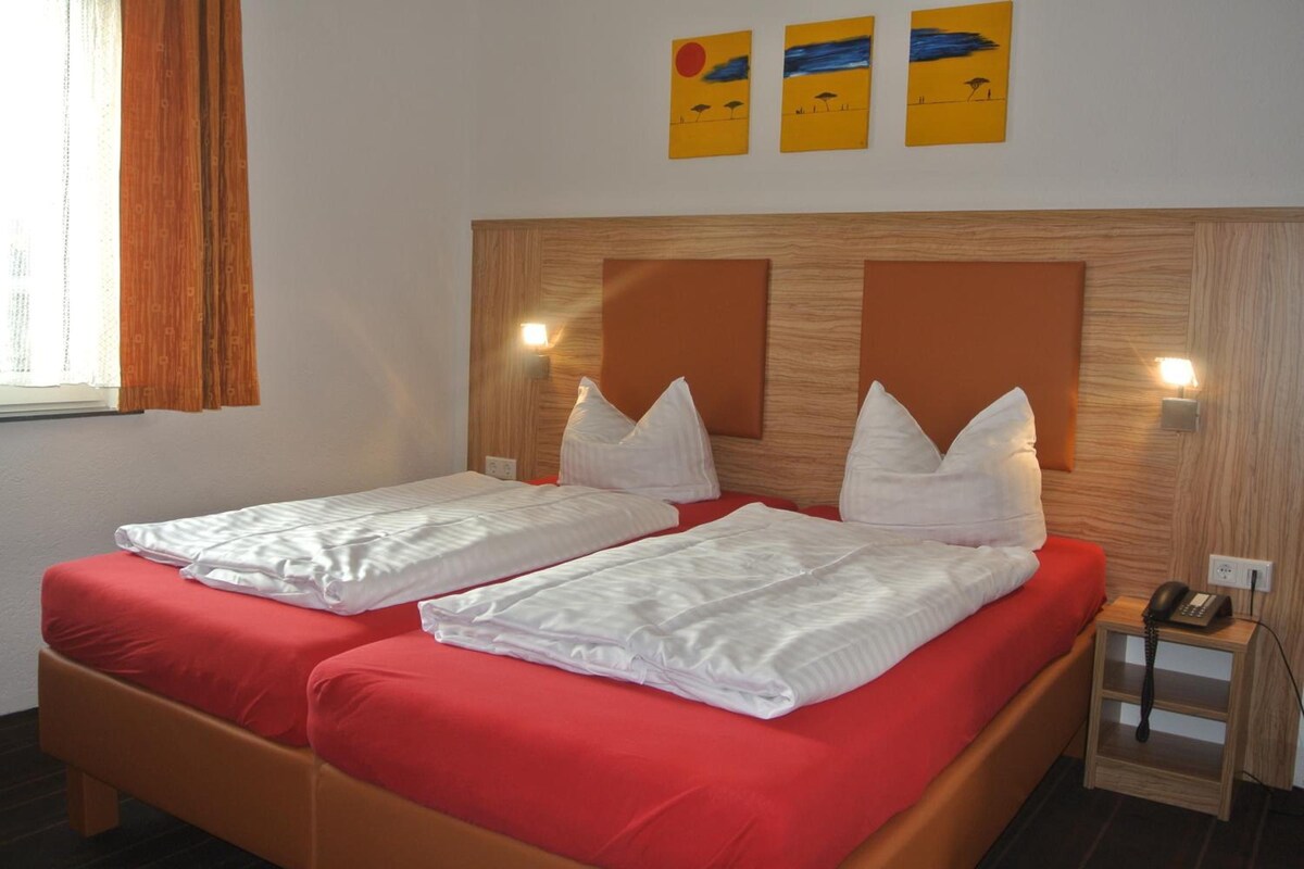 Habitación de hotel con dos camas individuales, ropa de cama roja, cabecero de madera, lámparas fijadas a la pared y tres cuadros sobre las camas.
