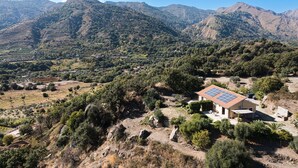 Une maison dotée d’un toit recouvert de panneaux solaires, entourée d’un paysage vallonné où l’on trouve des champs en terrasses et une végétation clairsemée.