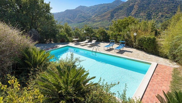 Une piscine rectangulaire à l’eau d’un bleu limpide, entourée d’une terrasse en pierre. Plusieurs chaises longues sont alignées le long du bord de la piscine. La piscine est mise en valeur par un décor de verdure luxuriante, avec des arbres et des collines en arrière-plan.