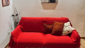 Un canapé rouge agr émenté d’un plaid et de deux coussins, un tableau abstrait encadré et un vase contenant des plantes séchées, le tout placé sur un mur.