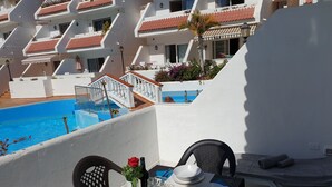 Un balcon avec vue sur la piscine, une table à manger avec des chaises et une petite table ornée d’un vase de fleurs.