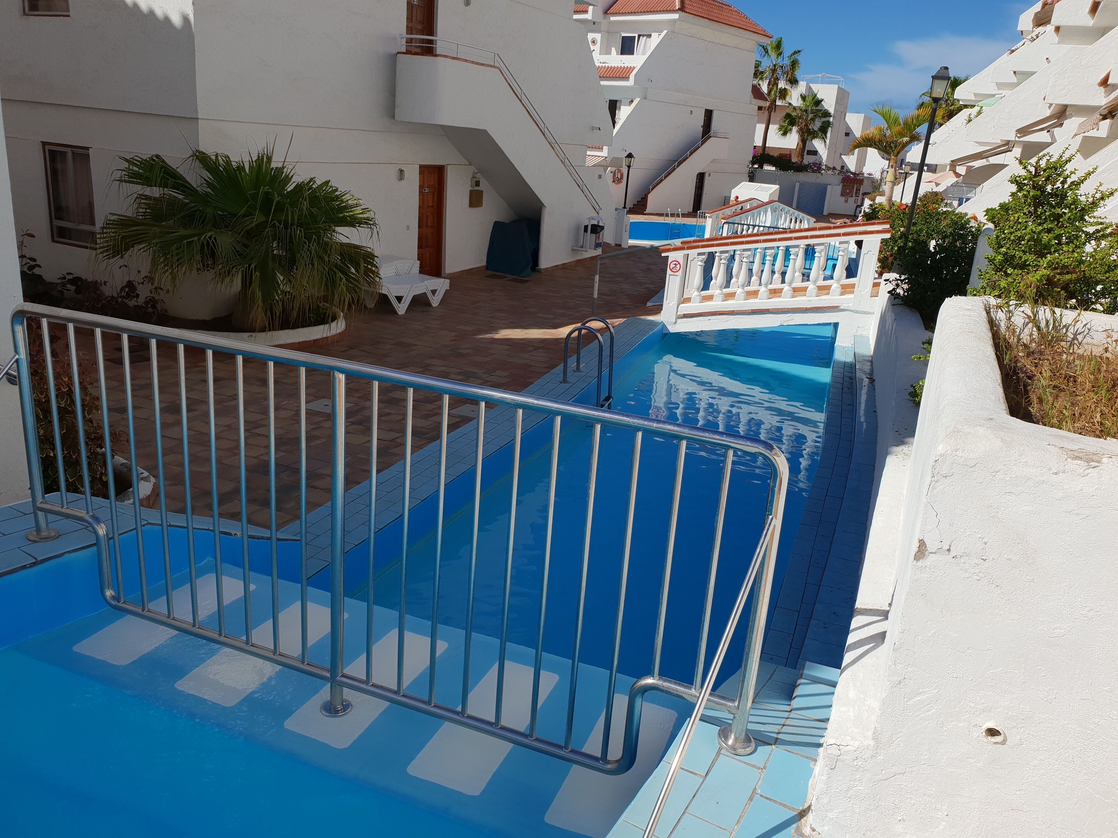 A swimming pool with a metal safety barrier, surrounded by white buildings with balconies and a clear blue sky.