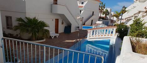 A swimming pool with a metal safety barrier, surrounded by white buildings with balconies and a clear blue sky.