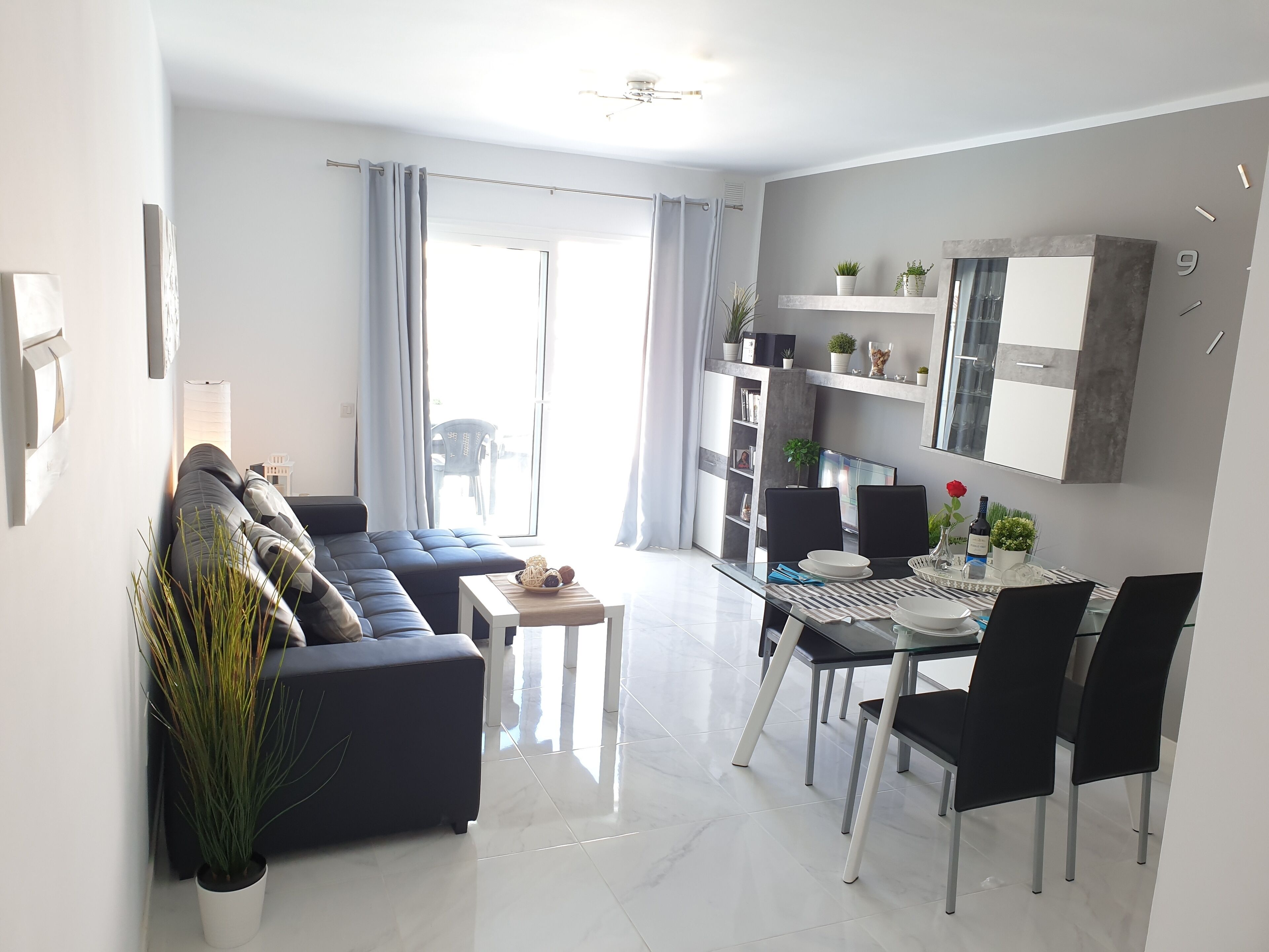 A modern living room with a sofa, dining table, chairs, and a small coffee table. There is a large window with curtains, a bookshelf, and a mounted mirror.