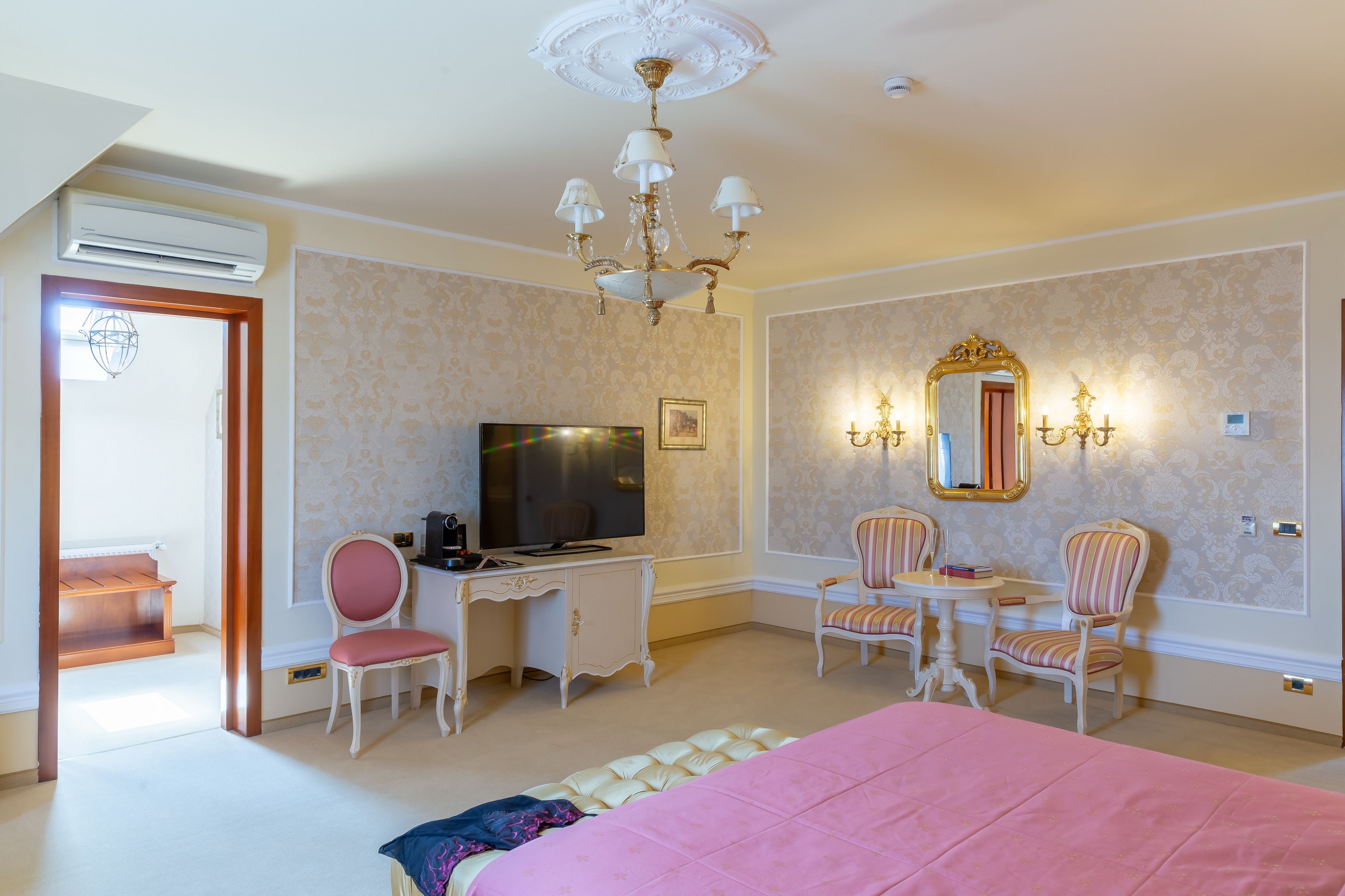 A hotel room with a bed, a desk with a TV, two chairs, a chandelier, and a large mirror.