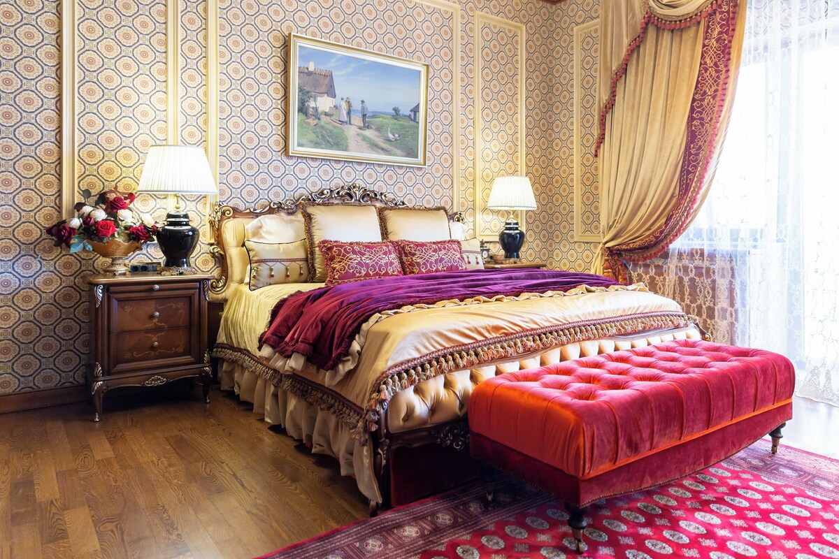 Une chambre à coucher classique, avec un grand lit, une tête de lit décorée, deux tables de chevet avec des lampes, un pouf rouge et un papier peint à motifs.