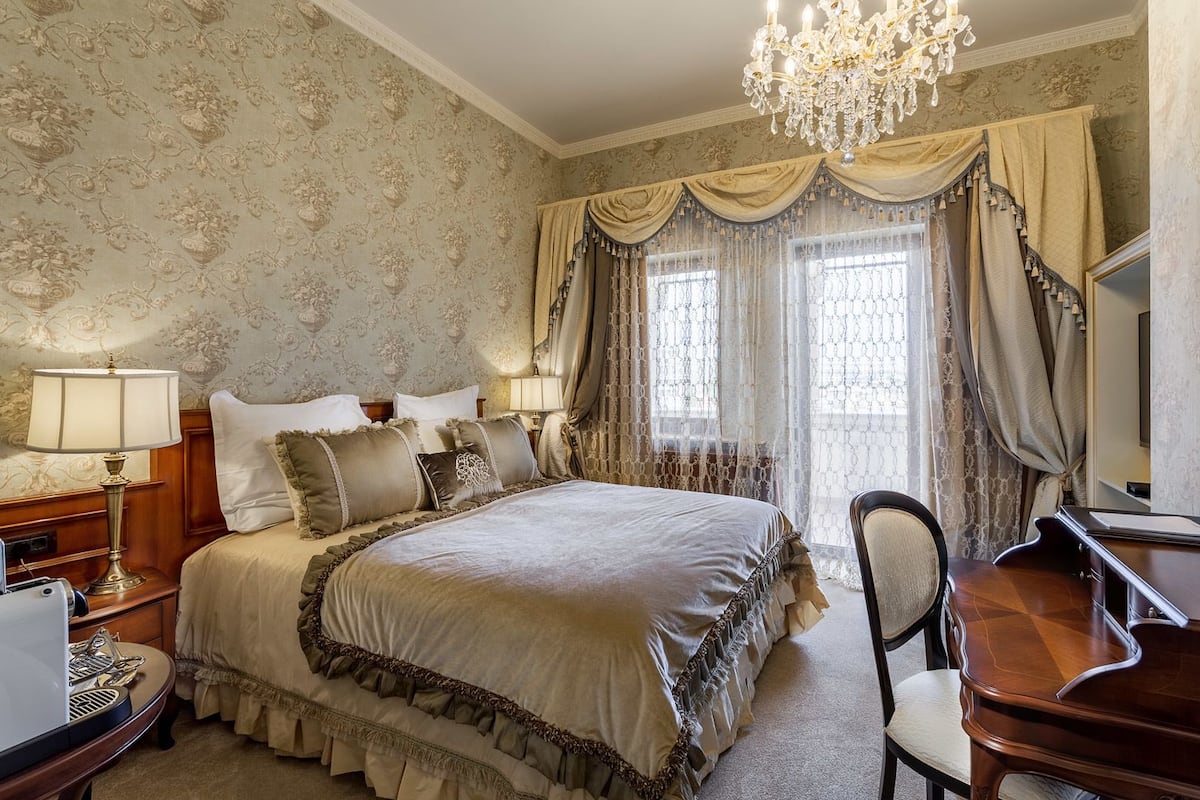 A classic bedroom with a large bed, ornate curtains, a chandelier, and a wooden desk.