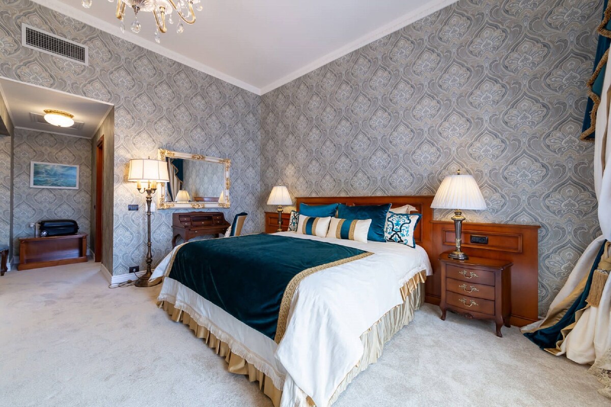 Une chambre spacieuse avec un grand lit, deux tables de chevet avec des lampes, une commode, un miroir et un lustre.