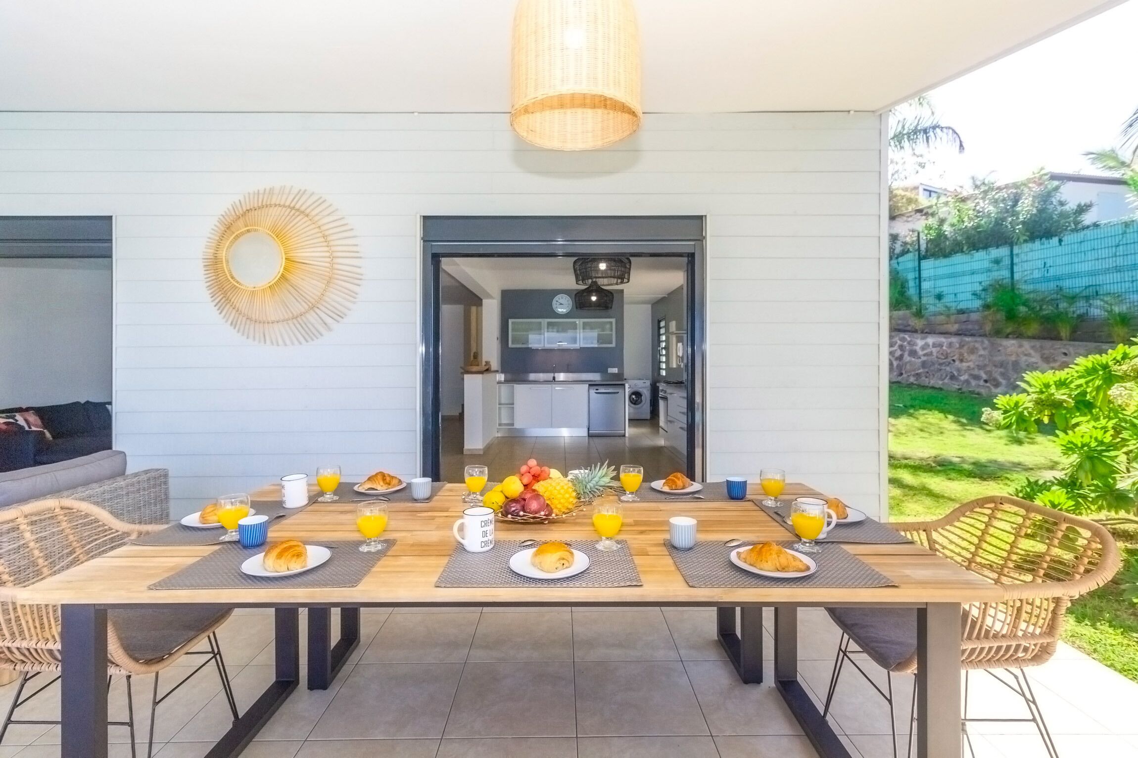A dining area with a wooden table set for breakfast, a kitchen visible through an open doorway, and a view of a garden.