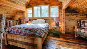 Une chambre confortable située sous les toits, avec un plafond en pente en bois, un grand lit recouvert d’une couverture à carreaux, deux lampes de chevet et un fauteuil agrémenté d’un coussin à motifs floraux.