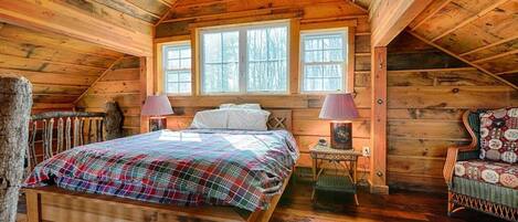 Un dormitorio acogedor en el ático con techo inclinado de madera, una cama grande con colcha escocesa, dos lámparas de noche y una silla con cojín floral.