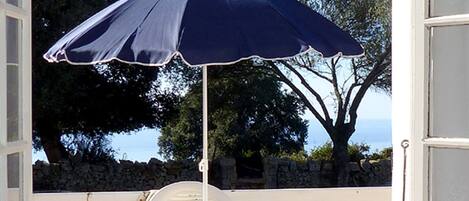 Een terras buiten met een tafel en stoelen, een blauwe parasol, en uitzicht op bomen en de zee.