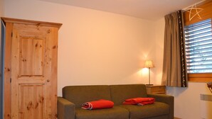 Ein Zimmer mit einem Holzkleiderschrank, einem grünen Sofa mit roten Decken, einem Nachttisch mit Lampe und einem Fenster mit Jalousien.