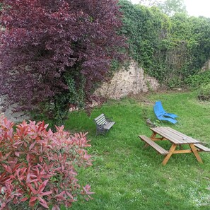 A grassy outdoor area with a wooden picnic table and blue chairs, surrounded by bushes and a stone wall. - Pretty cottage F2 of 50m ² independent in Rouffach (Rouffach)