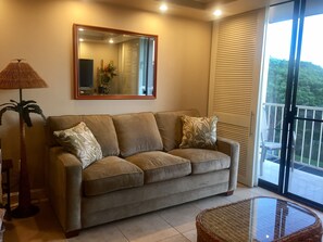 A living room with a sofa, a glass coffee table, a floor lamp, and a large window with blinds.