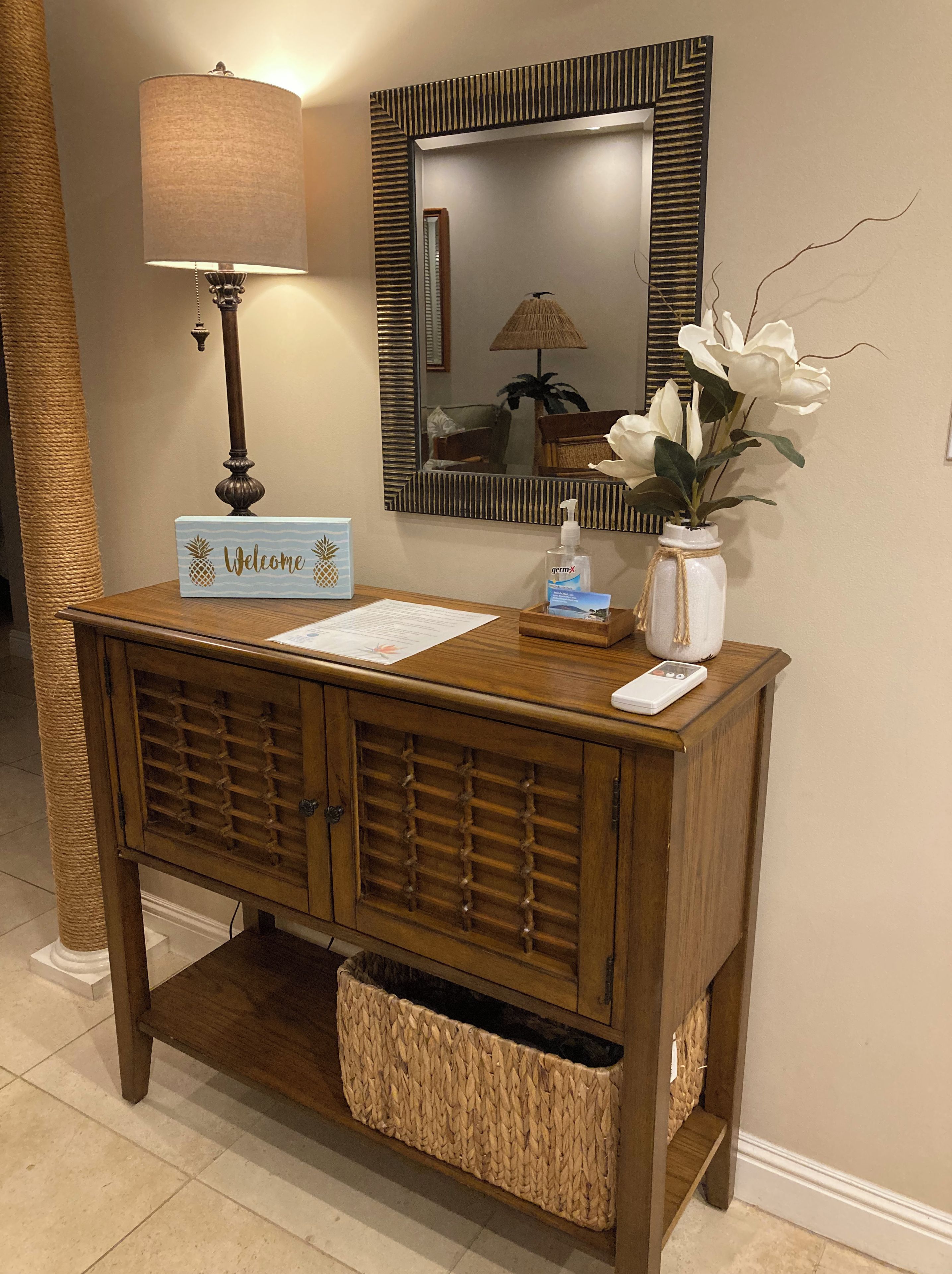 A wooden console table with a 'Welcome' sign, a lamp, a vase with white flowers, and a tissue dispenser.