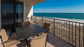 A balcony with a table and chairs, overlooking a beach with the ocean and a clear sky.