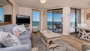 A living room with a TV, grey sofa, and a wooden coffee table. There is a balcony with a view of the beach. - 3BR 2BA **BEACH FRONT!** Phoenix 7 **Rsvd Parking Space! (Orange Beach)