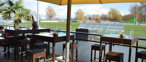 An outdoor patio with tables and chairs, a large umbrella, and a view of a sports field.
