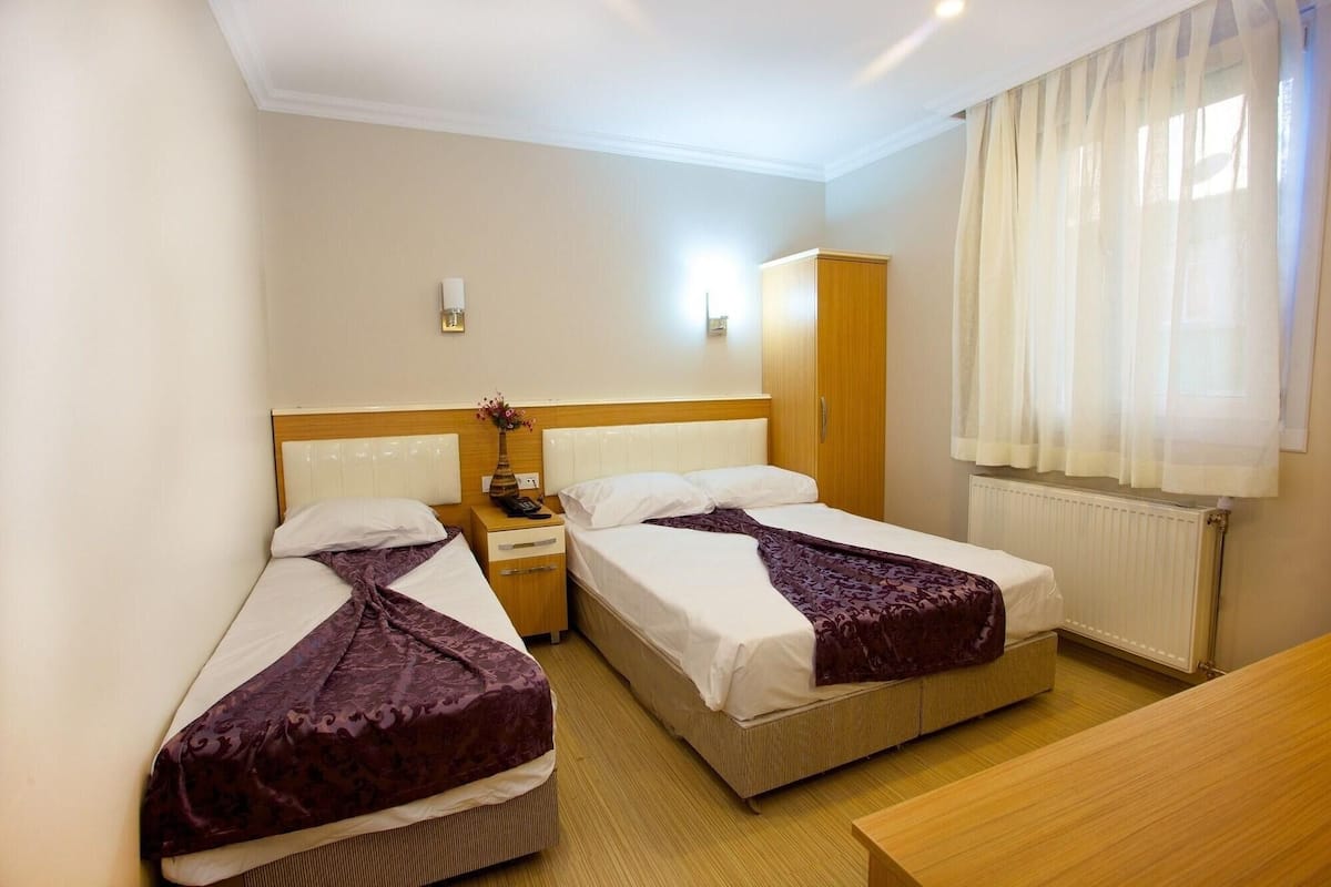 A hotel room with two beds, a wooden nightstand, a wooden wardrobe, and a window with sheer curtains.