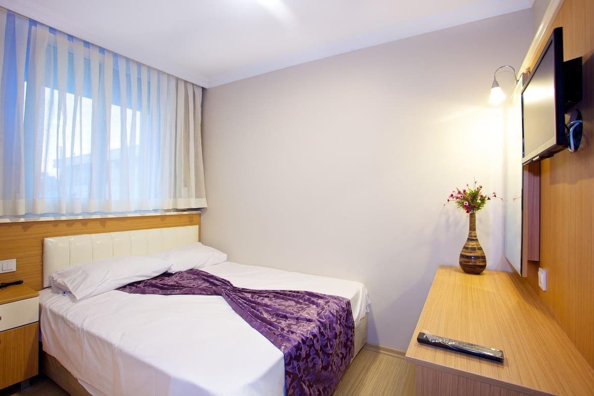 A hotel room with a bed, a nightstand, a wooden desk, a vase with flowers, and a television mounted on the wall.