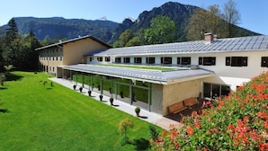 A modern building with a covered walkway, a lawn, and mountains in the background. - BSW Hotel Hubertuspark (Schoenau am Koenigssee)