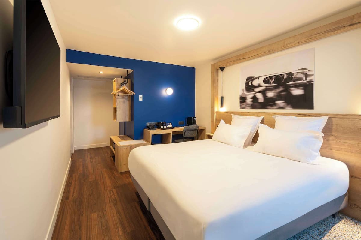 A hotel room with a large bed, a desk with a chair, a wardrobe, and a mounted TV.