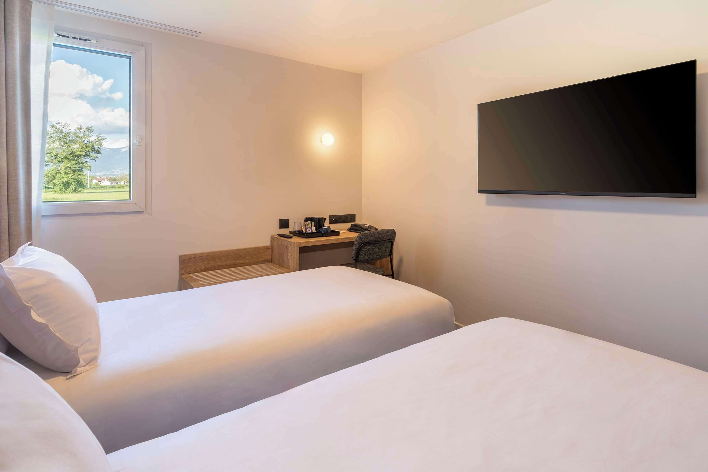 A hotel room with two beds, a desk with a chair, a flat-screen TV mounted on the wall, and a window with a view of greenery and buildings.
